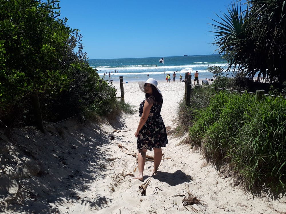 chica morena con un vestido de flores y un sombrero blanco girada mirando a cámara sonriente. Enfrente de ella hay la playa